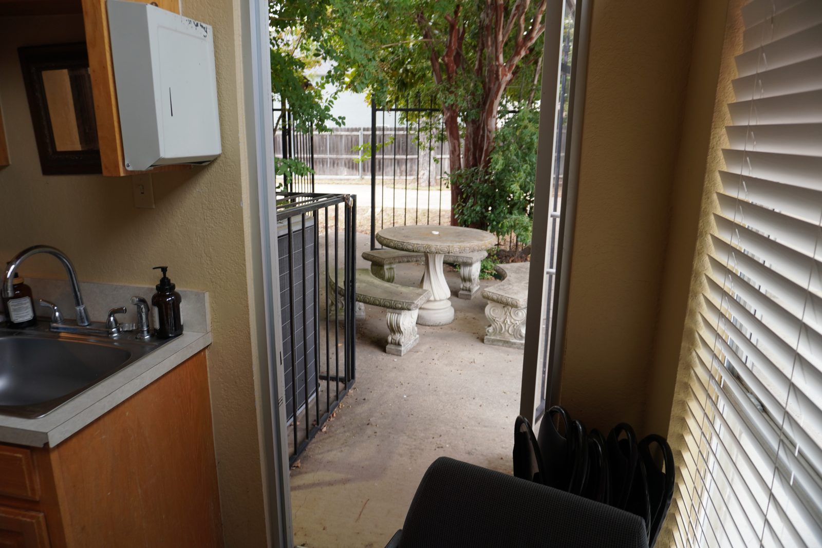 Open glass-panel door looking out to a paved courtyard with a stone bistro table and mature trees.