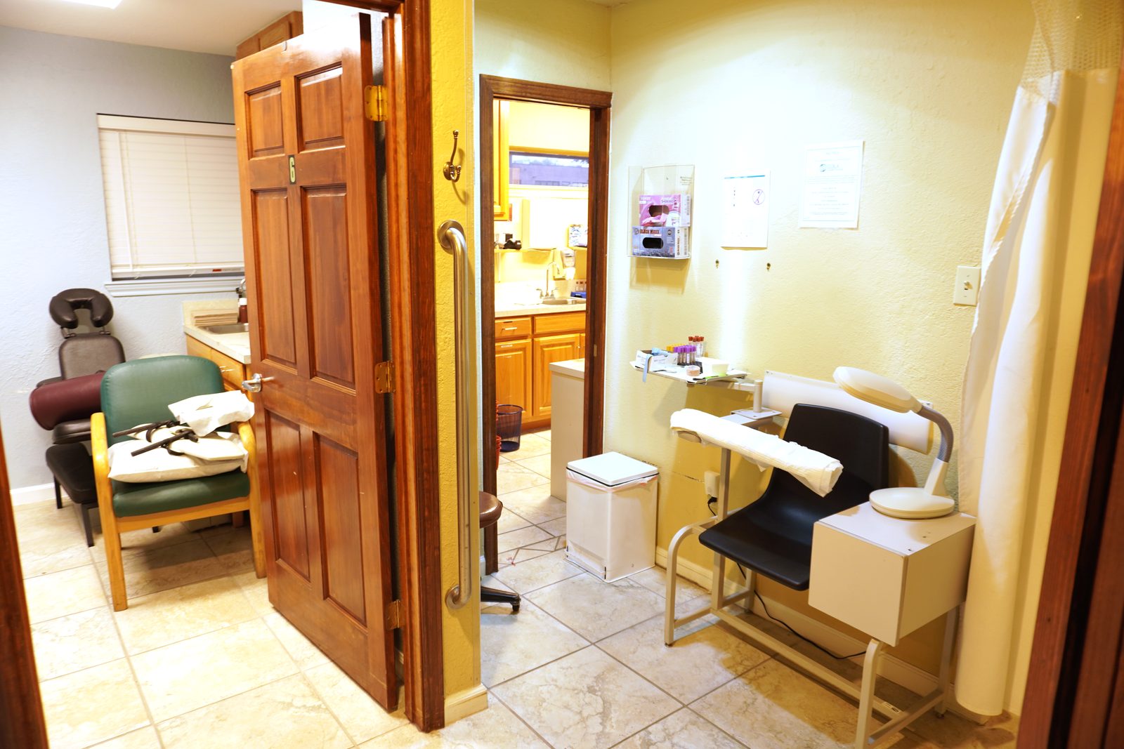 Examination room with a treatment chair and paper roll, opening onto a hand-wash alcove through a honey-oak door.